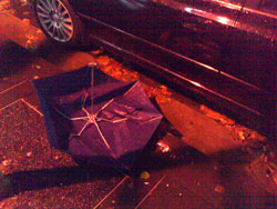 Photograph of a broken umbrella, lying next to a car on a dark street.