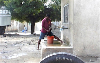 Photograph or rural Tanzanian village, with man using hand pump to get water