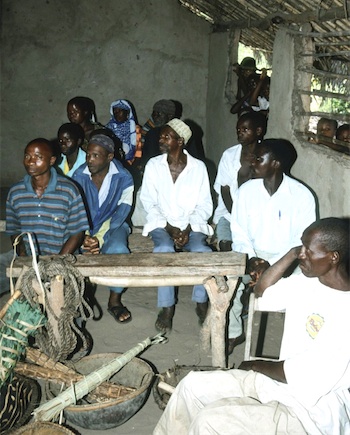 Photograph of Tanzanian villagers meeting in a hut with a rough-hewn wooden table and basic household utensils