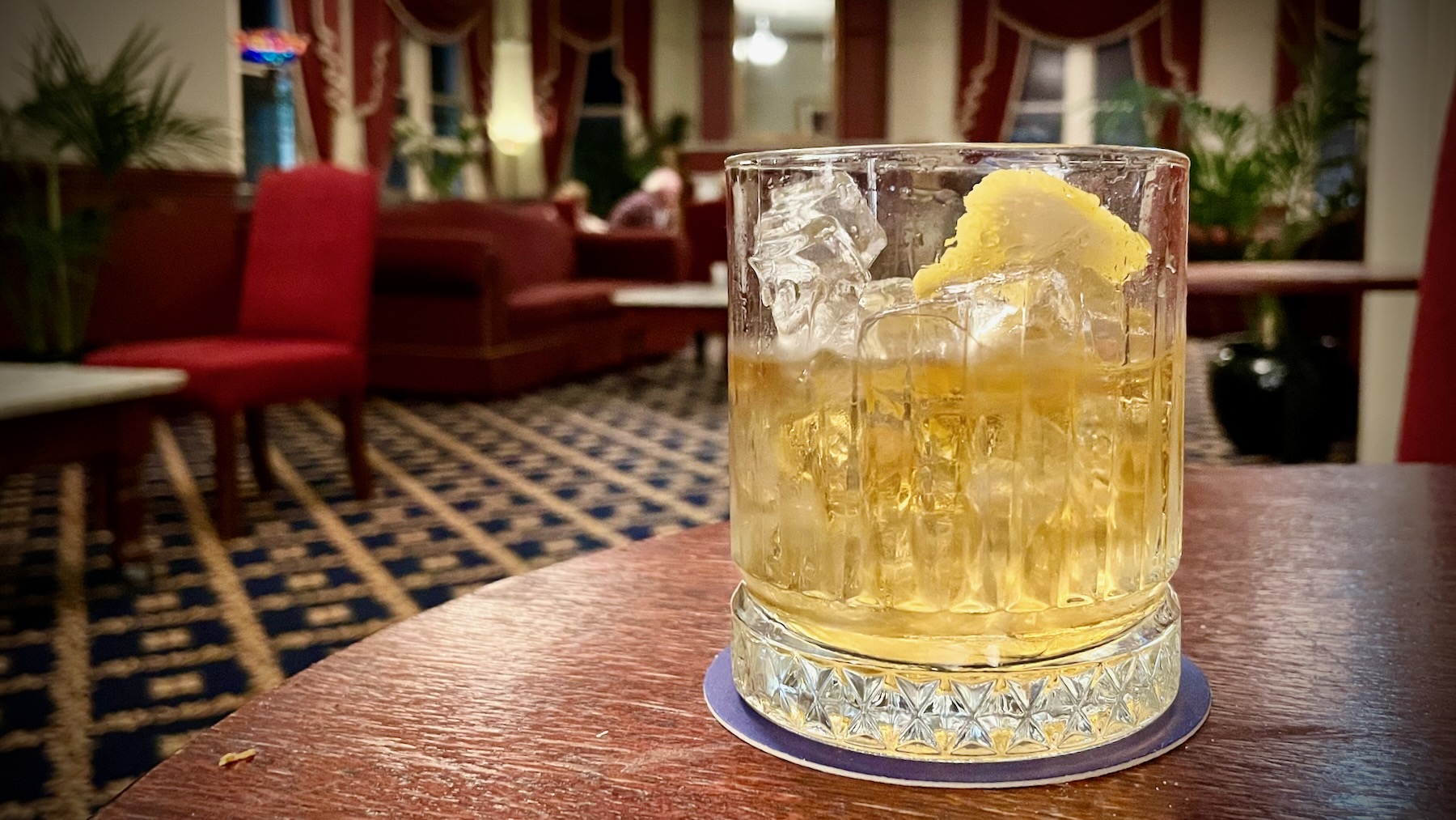 On an old wooden table sits an old-style lowball glass containing a pale yellow liquid, ice cubes, and slice of lemon. In the background, out of focus, the lush dark red furnishings of a high-Victorian drawing room.