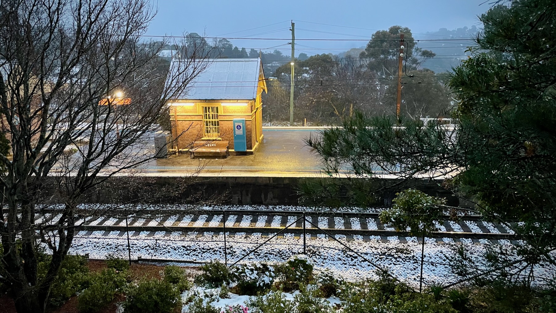 A view downhill in the evening towards a Victorian-era railway station, or at least its equipment hut. The platform is lit up with an orange tinge, but the sky is grey, and snow is scattered along the track.