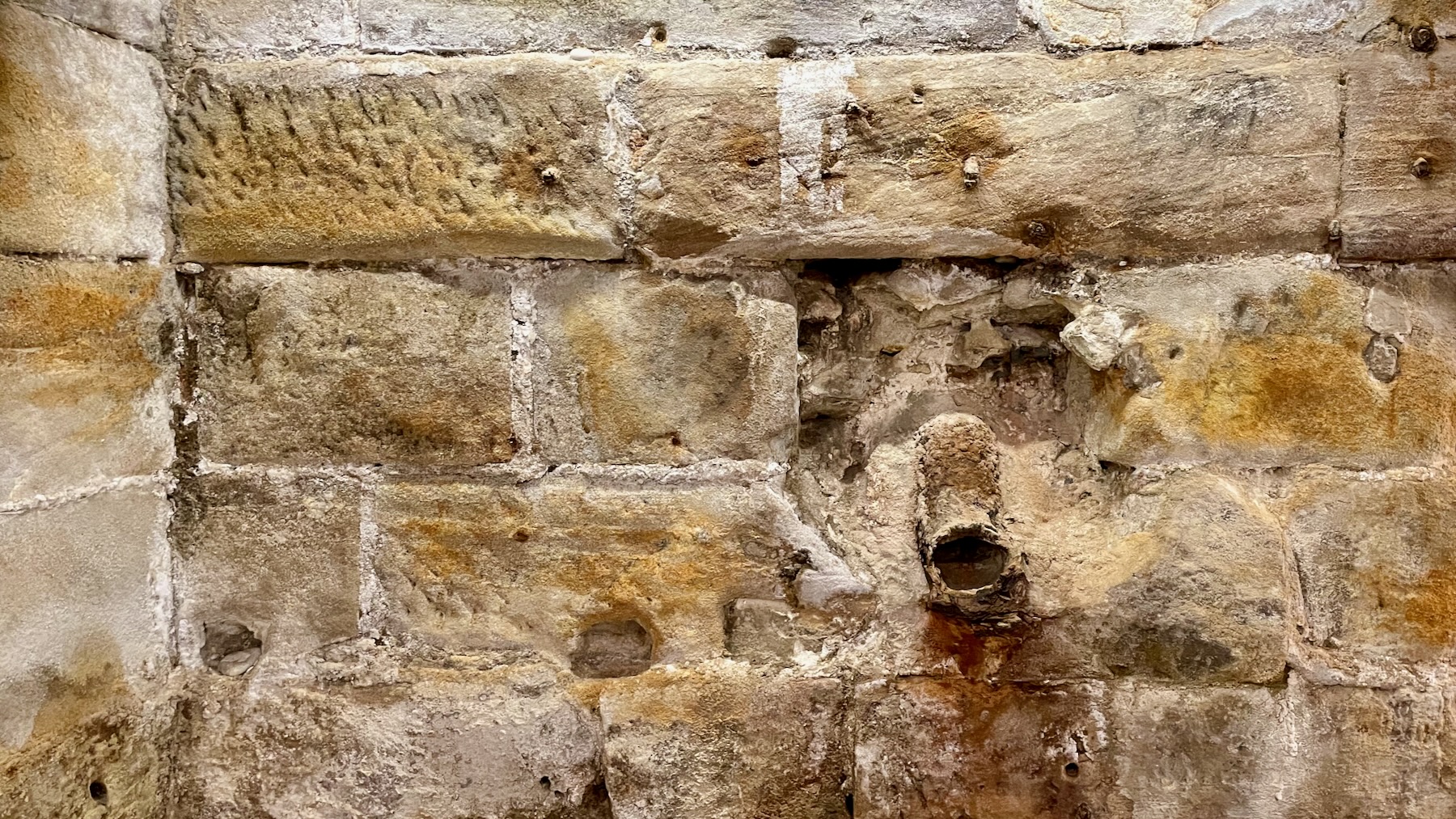 Photograph of a colonial-era sandstone wall, being the foundations of the Paragon Hotel at Circular Quay. On the lower right of the image is the rusty remnant of a pipe and spout entering the room. The beauty is in the textures.
