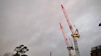 Against a grey cloudy sky are highlighted two massive construction cranes, their bright colours highlighted by the setting sun.