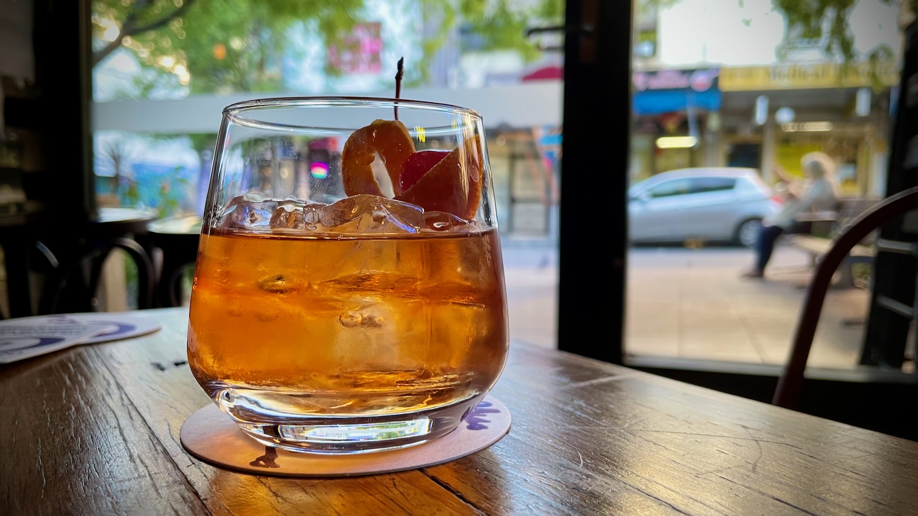 A simple lowball glass containing a tan-brown liquid and ice cubes, a twist of orange, and a maraschino cherry. And in the background, through a window and out of focus, a quiet suburban street in the last, late afternoon.