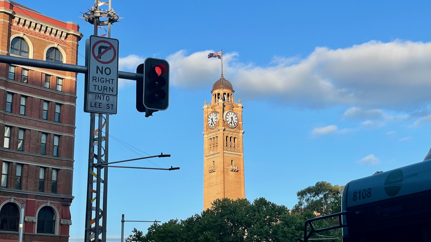 In the middle distance, the late afternoon sunlight catches a sandstone clock tower which rises above the treeline. The view is framed on the left by a red brick building and a modern stainless steel lighting tower on which there's a traffic light (red) and a NO RIGHT TURN sign, and on the right by the silhouette of a bus.