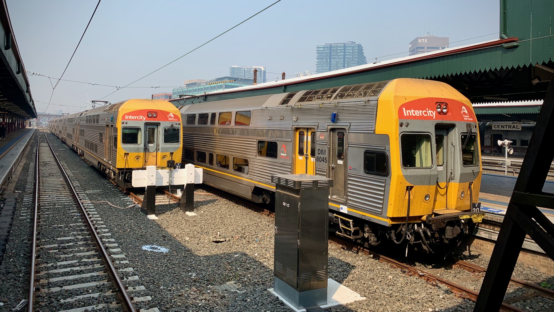 Looking from the concourse end of Sydney Central terminal down the tracks between two platforms. Sitting at the right-hand platform is a V-Set train, with its fluted stainless steel body and bright yellow and red nose. On the centre stabling track is another. There’s nothing at the left-hand platform. Overhead, it’s a sunny sky smudged with the brown of bushfire smoke.