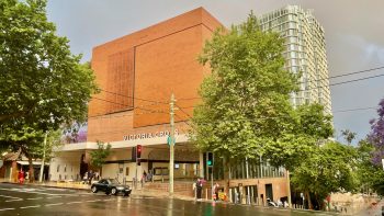 A view across a street wet from recent rain to a vast brick block, which hides the ventilation shaft for a Metro station. Along the side, on the verandah overhanding the entrance stairs, are the words VICTORIA CROSS. Nearby, a healthy-looking London plane tree.