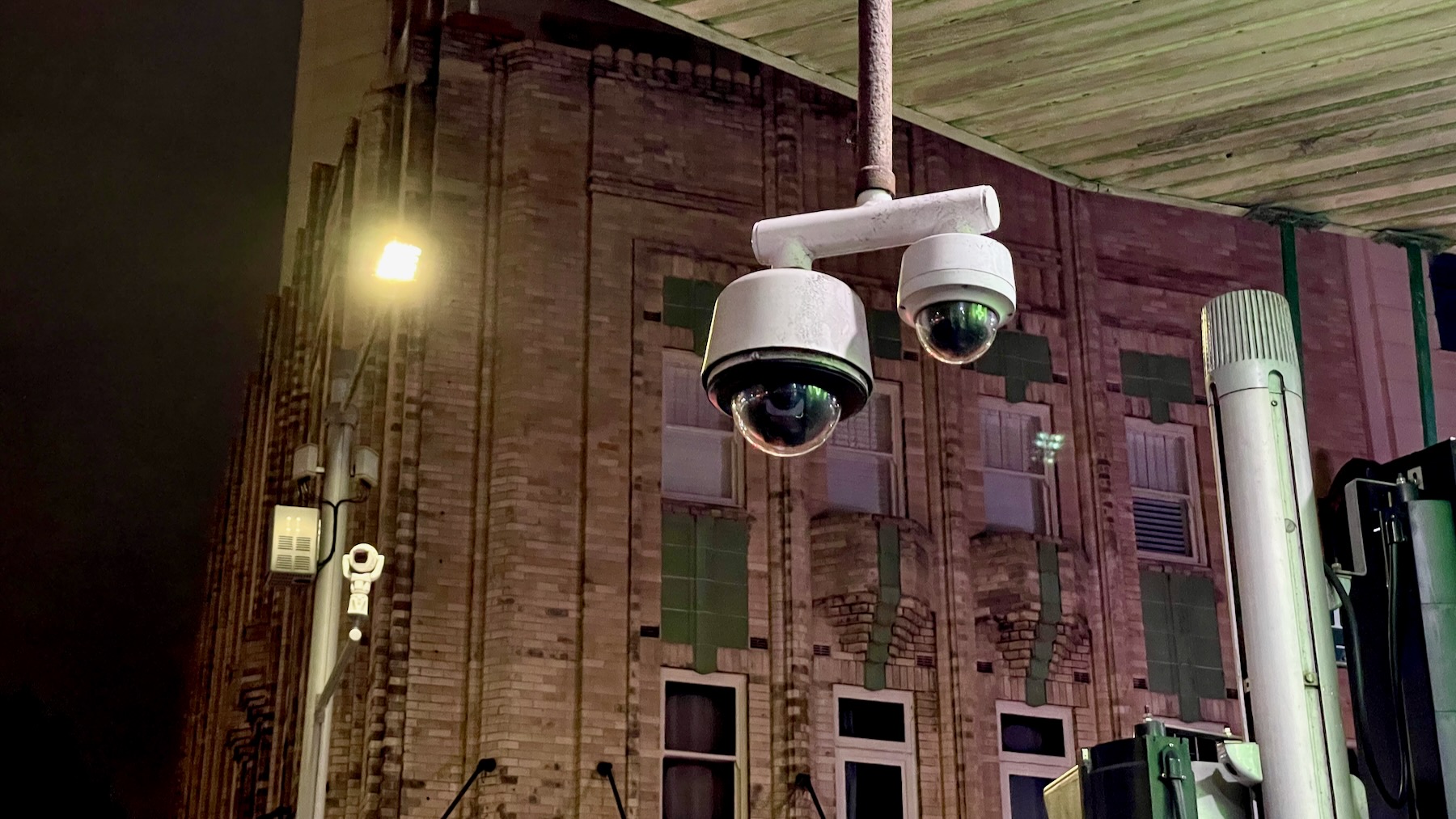 Nighttime, looking from a city footpath up towards a pair of surveillance cameras in their black domes under white bodies. Across the street are the upper floors of an art deco brick office building, in front of which is another surveillance camera, this one steerable.