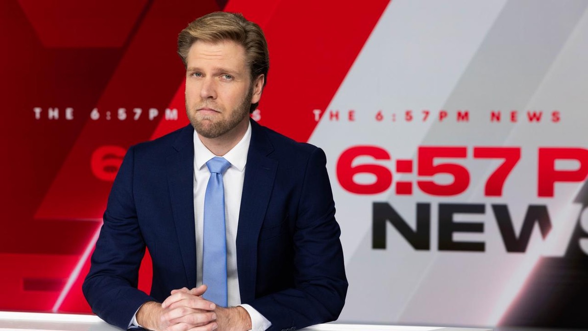 A white man in a dark suit, white shirt, and pale blue tie sits with his hands folded at the news desk. He is tall, blond, and neatly bearded, He looks sad.