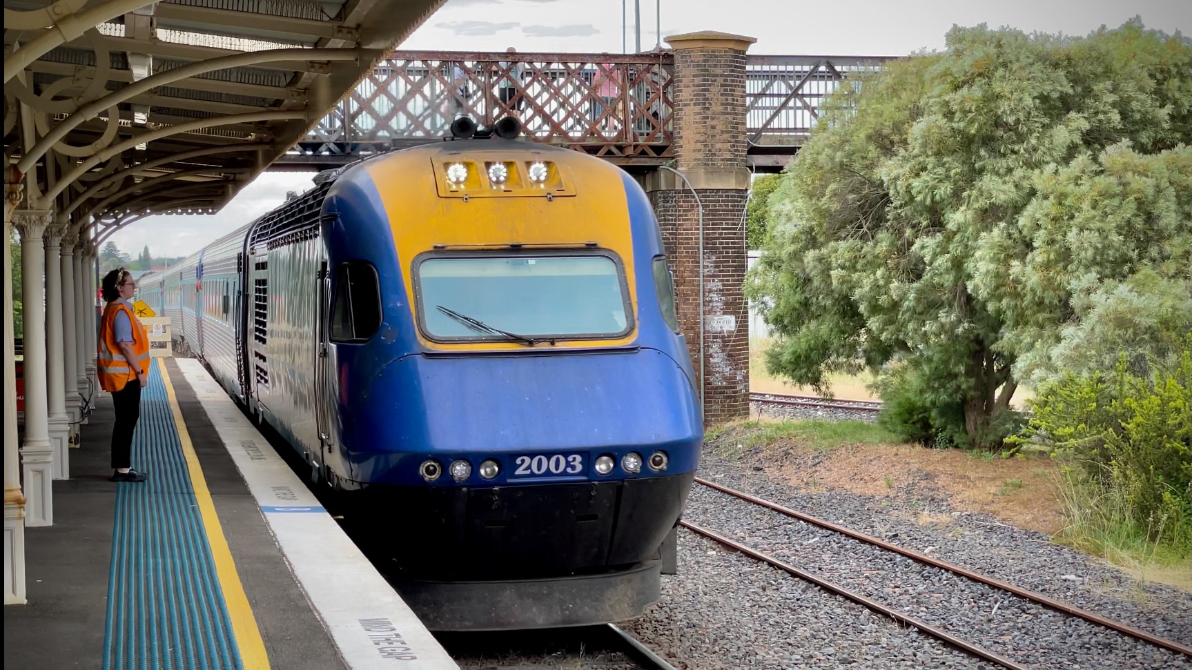 Photo of a NSW TrainLink XPT power car number 2003 and its train on the platform at Orange station. Its sloped front has a single full-width window for the driver, and it’s painted blue with a yellow panel around the window and the three headlights at the top.