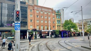 A view across a city intersection on a rainy day. In the centre of the frame is the Great Southern Hotel, Sydney, a five-storey red-brick Art Deco construction nestled between an modern glass office building and a narrow Victorian-era four-storey business building. Two tram tracks run from behind the viewer through the intersection and to the right.