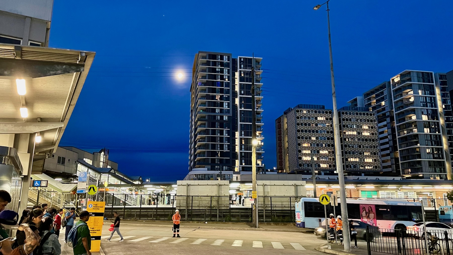 In the middle distance, across a floodlit concrete area, is a brightly lit suburban railway station. On the left, people are waiting for rail replacement buses. A temporary yellow sign with black lettering lists the destinations. In the centre, a man in hi-viz and a white safety helmet stands watching it all, his hands clasped behind his back. In the distance, cross the tracks and behind the station, stand several modern apartment buildings. The tallest rises around 20 storeys, and to its left against the deep blue dusk sky is the Moon, three days from full.