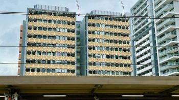 Seen across the roof of a suburban railway station are two modern apartment buildings their facades decorated with hexagonal shapes in matt gold with white trim.