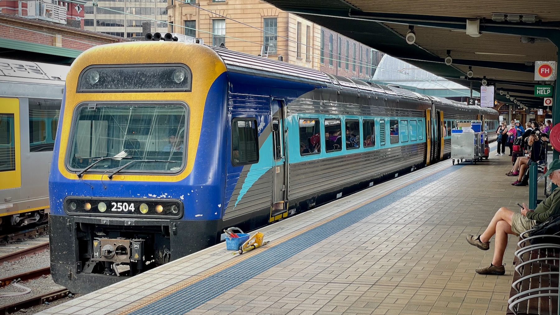 A NSW TrainLink Xplorer three-car DMU stands at Sydney Central station. A single deck train in silver with a light blue stripe along the window line, dark blue nose with a bright yellow highlight around the front window and upper lights, and dirt all over. It’s numbered 2504. On the platform sit some cleaning products, clearly only used on the inside of the trains, and passengers sit waiting for the cleaning to be finished.