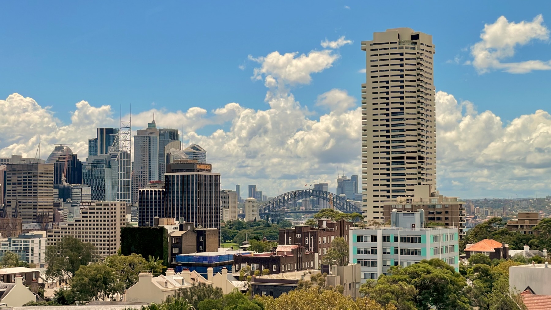 A view from the 13th floor across Darlinghurst to the north-west. On the left are the towers of the northern end of the Sydney CBD. In the middle, in the distance, one can see the Sydney Harbour Bridge, and on the right the view is dominated by Harry Seidler’s Horizon Apartments, a white modernist tower with an grid-based structure. The day is bright and sunny with a few fluffy white clouds.