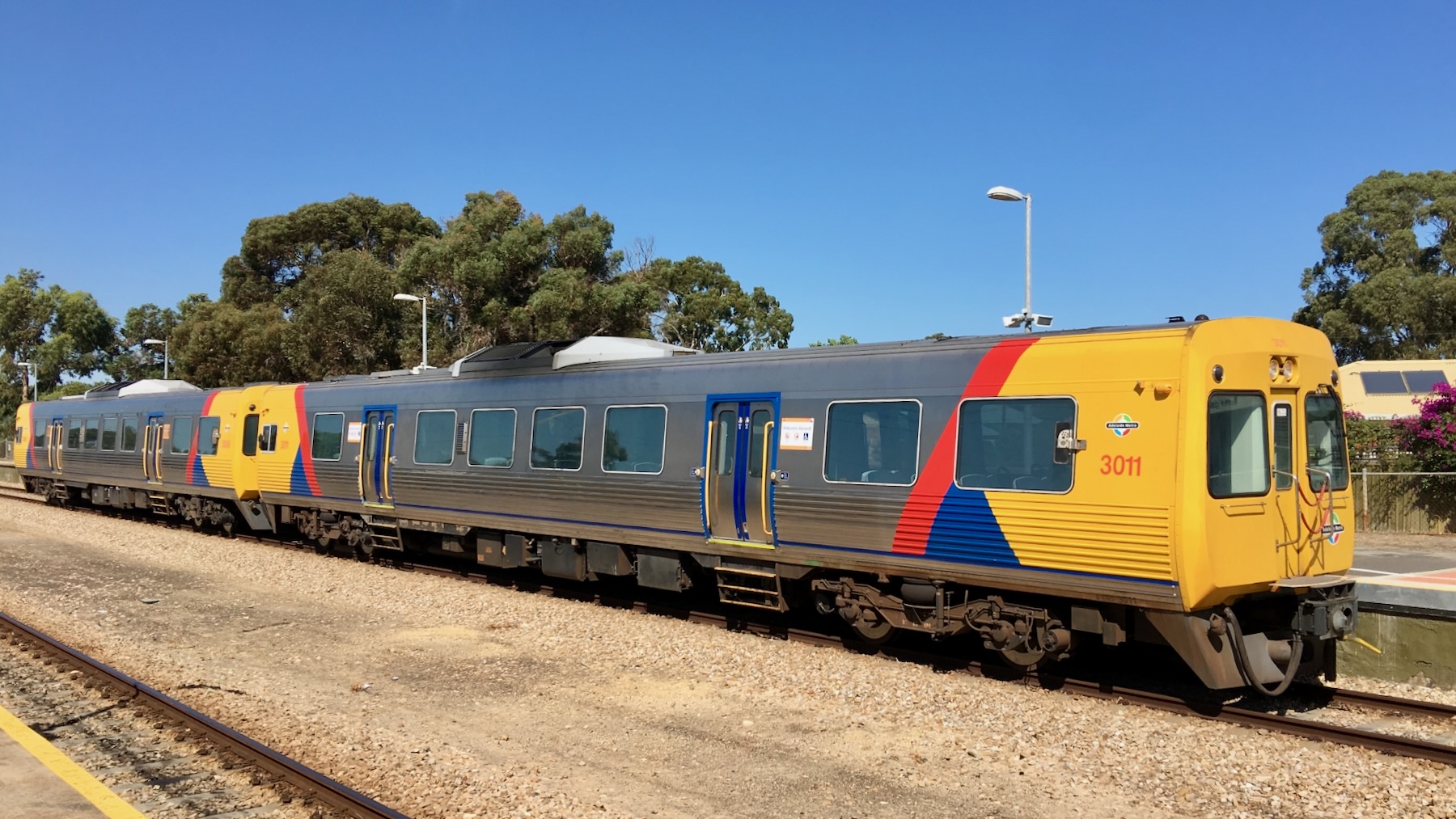 On a bright sunny day a pair of diesel railcars sit at a station platform in the open air. They’re silver, with their flat noses painted bright yellow, and just behind that a red stripe and a a small flash of blue.