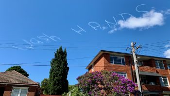 In the blue sky above a typical inner west Sydney red-brick block of flats a skywriter has scrawled WASH HANDS.