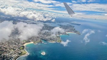 The view from the left-hand window of an airliner over the eastern beaches of Sydney. About two-thirds of the way up the view is Sydney Harbour in all its crinkly glory. Along the lower parts of the image are the beaches, with Bondi uppermost. Near the top of the image a bank of white fluffy clouds obscure the North Shore. And at the very top right, the aircraft’s wing.