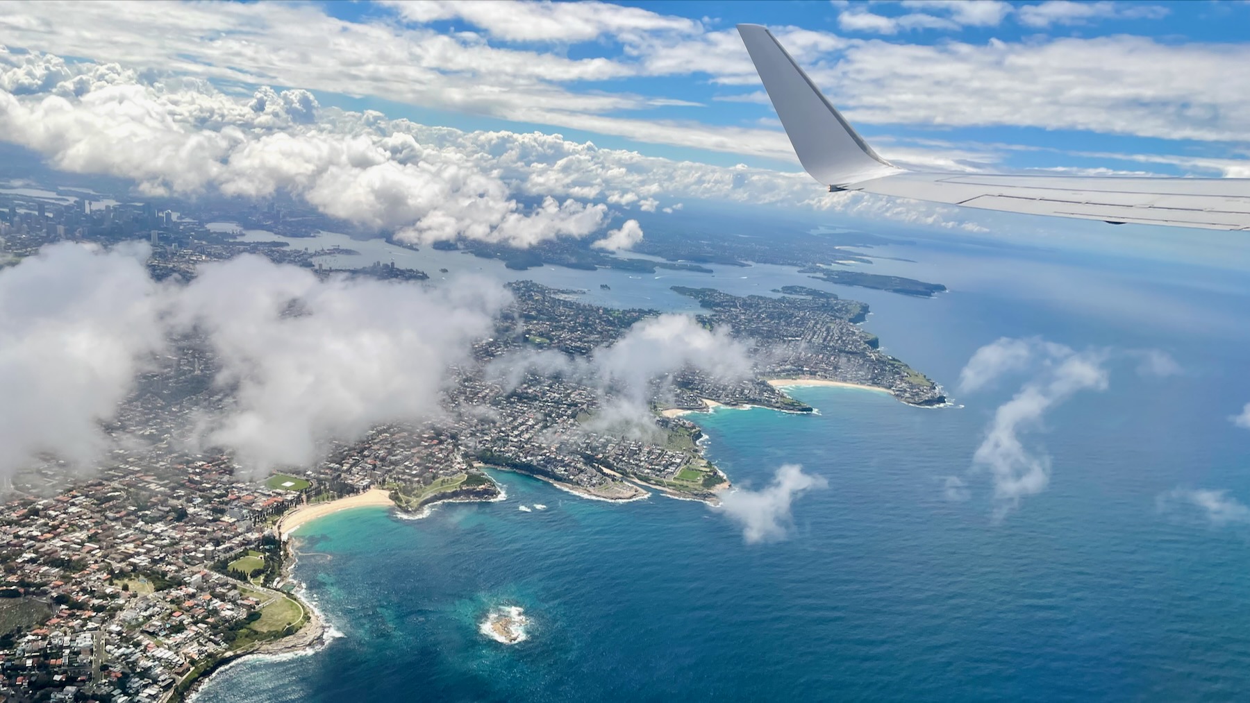 The view from the left-hand window of an airliner over the eastern beaches of Sydney. About two-thirds of the way up the view is Sydney Harbour in all its crinkly glory. Along the lower parts of the image are the beaches, with Bondi uppermost. Near the top of the image a bank of white fluffy clouds obscure the North Shore. And at the very top right, the aircraft’s wing.