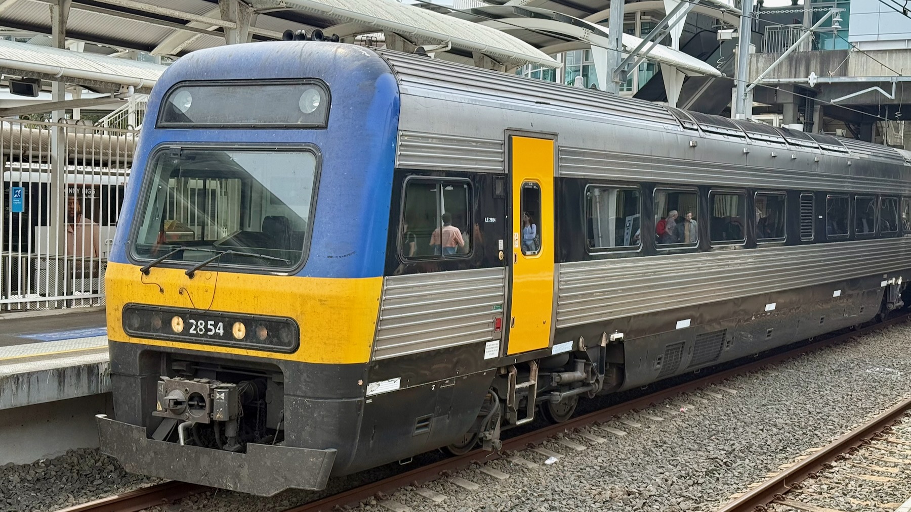A NSW TrainLink Endeavour railcar at Blacktown station. It’s a diesel multiple unit (DMU) although we can only see the first of two cars, facing to the left. It’s made of fluted silver metal sides with a black band running along the windows. The front end is painted dark blue with a bright yellow band below the driver’s window, and it has bright yellow doors. The underparts and black, and the entire train is filthy and in need of a good wash. Its number is 2854.