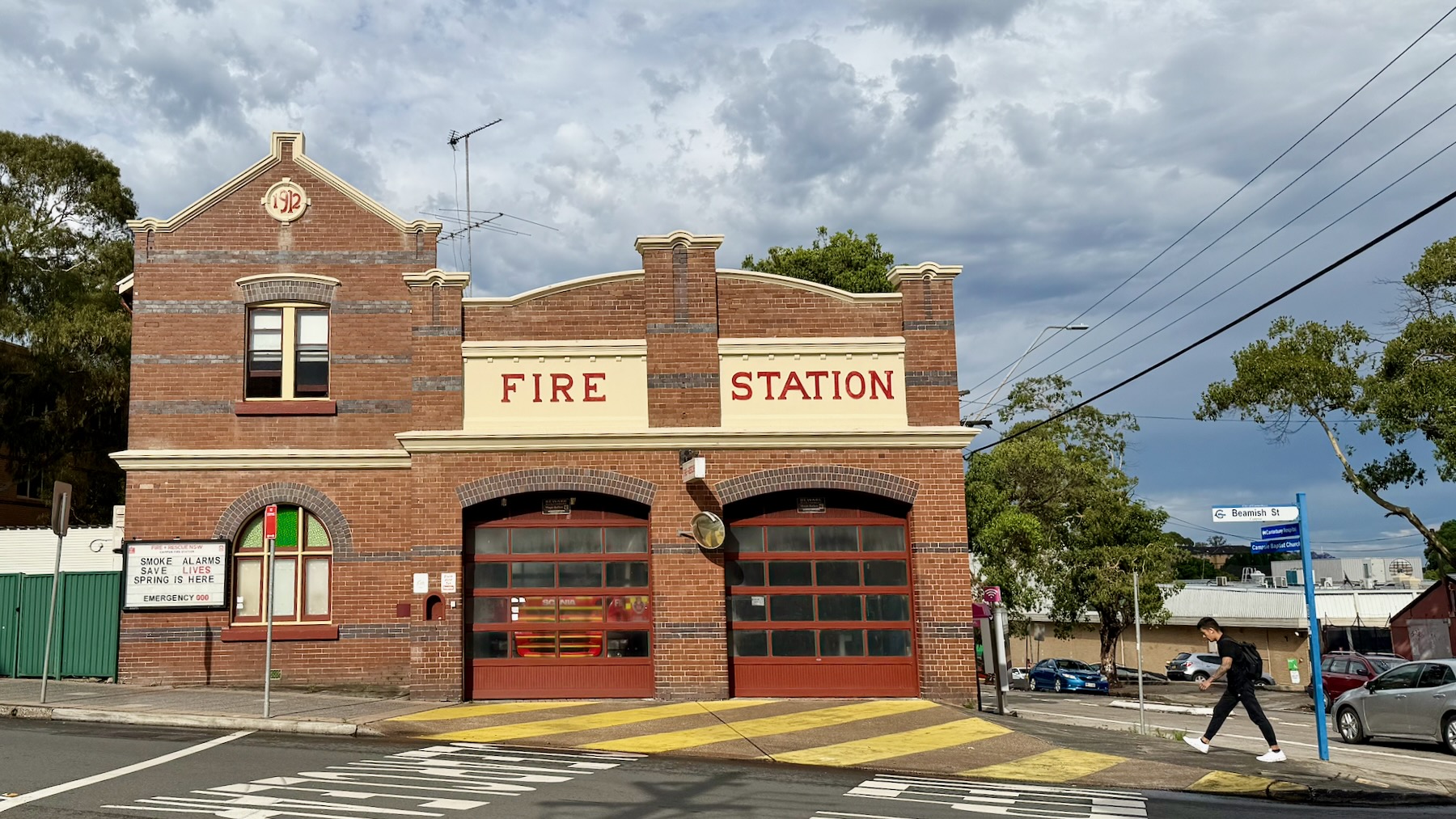 A suburban street in Sydney’s inner south-west. A two-storey red-brick building is labelled “1912” and ”Fire Station” in early 20th century typography in maroon on cream. Two big garage doors hide a modern fire engine and an empty garage respectively. A sign on the lower left reads: “Smoke alarms save lives. Spring is here. Emergency 000.” On the right, a young man wearing lack strides into the frame. Overhead, rainclouds loom.