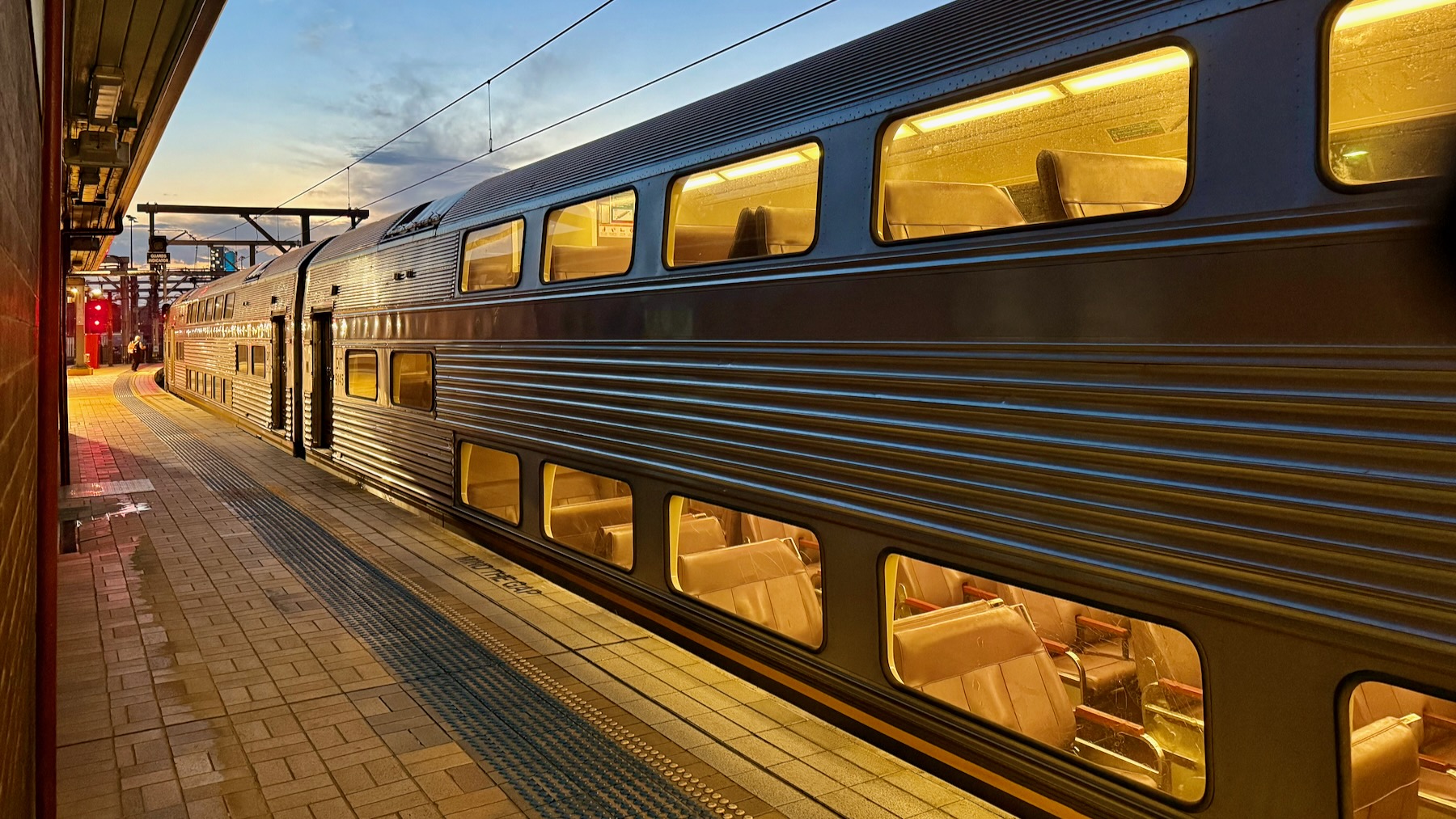 Looking along a railway station platform shortly before sunset. A Sydney Trains V-Set stands on the right, stretching along the platform towards left, where its distant nose is just short of a signal glowing red over red. The forward part of its shiny fluted steel sides glisten in under the platform’s illumination, although the car nearest us is mostly in the shadows, its two levels of windows glowing yellow and highlighting the purple upholstery within, The only person to be seen is the driver walking up to the front of the train, a tiny figure in the distance.