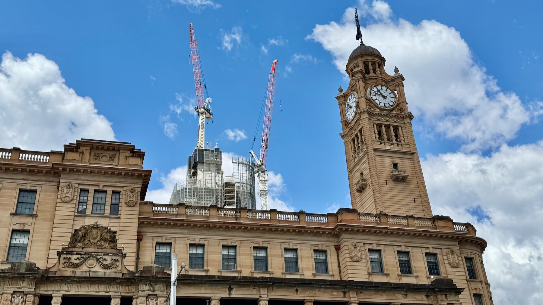 The upper parts of the sandstone facade of Sydney Central station, including the coat of arms on the left and the clock tower on the right. Behind it are two construction cranes and the partly-constructed modern architecture of Atlassian Central. And behind all that is a bright blue sky with fluffy white clouds.