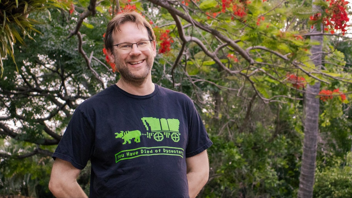 A middle-aged white man in a black t-shirt stands in front of some tropical tree or other, smiling, with his hands clasped behind his back. His hair is brown, as is his beard. He wears spectacles . The t-shirt has a green print in the style of, and indeed taken from, an old video game called Oregon Trail. It shows a covered wagon and the text: You Have Died of Dysentery.