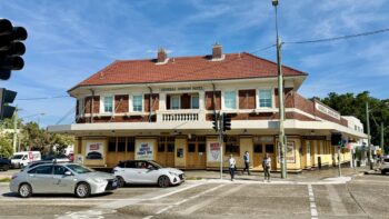 An art deco pub on a bright sunny day. Its ground floor has floor to ceiling dark cream tiles with brown outlines and a series of wooden doors. The upper storey is red brick with light cream window frames. The roof is red tiles.