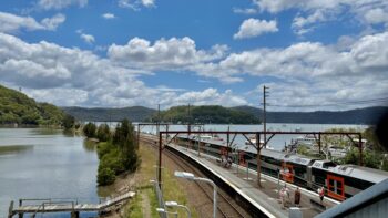 A view across a large estuary to a tree-covered island and then to the forested banks beyond on a bright sunny day. In the foreground on the right is a railway station platform, and on the other side a Sydney Trains D-Set Mariyung train stands, having just disgorged some passengers. The twin railway tracks sweep from the right into the centre and middle distance, along a causeway, to an island and into a tunnel.