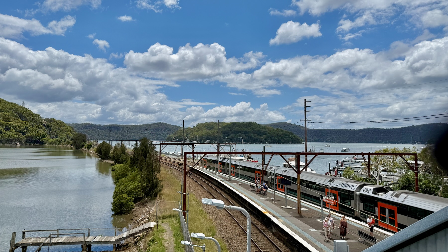 A view across a large estuary to a tree-covered island and then to the forested banks beyond on a bright sunny day. In the foreground on the right is a railway station platform, and on the other side a Sydney Trains D-Set Mariyung train stands, having just disgorged some passengers. The twin railway tracks sweep from the right into the centre and middle distance, along a causeway, to an island and into a tunnel.
