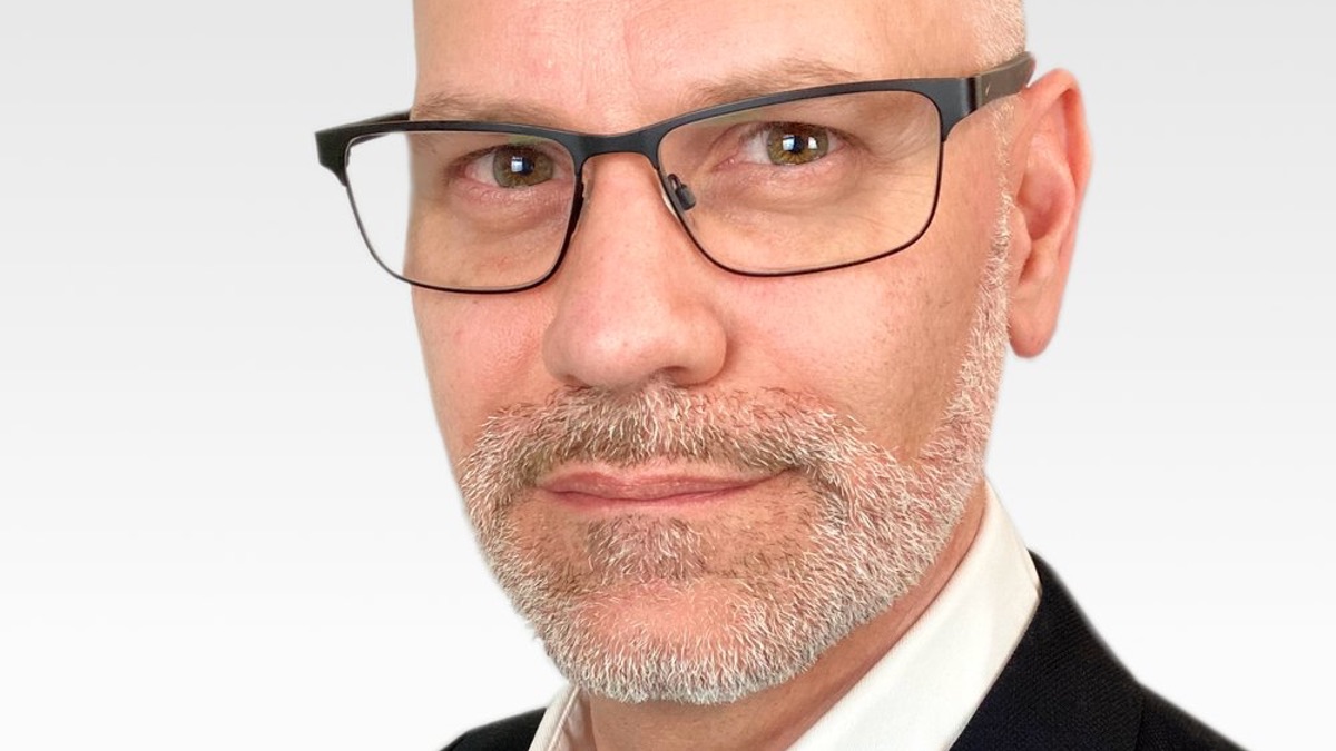 Close-up photo of middle-aged white man with neatly-trimmed grey stubble and black-rimmed spectacles. He wears an open-necked white collared shirt and a black jacket