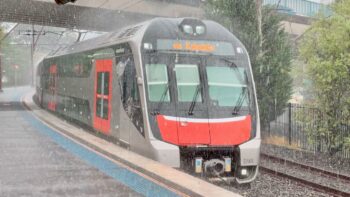 A Sydney Trains D-Set Mariyung train arrives at Katoomba station in the pouring rain. It's grey with black bands along the windows of its two decks and safety orange doors. Its blunt nose is black with light grey trim and a big chunk of safety orange below the driver's cab.