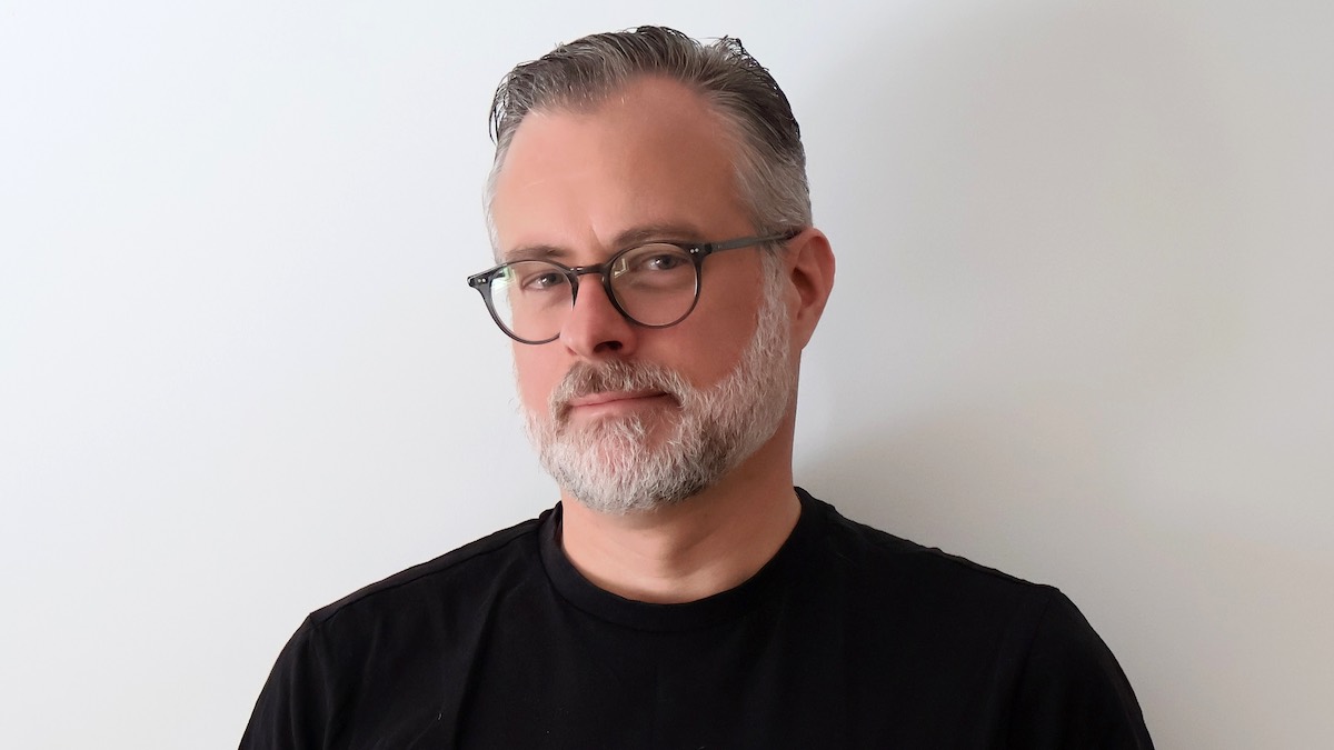 A middle-aged white man with a greying beard and neat black spectacles wears a black collarless top. He looks at the camera with thoughtful scepticism.