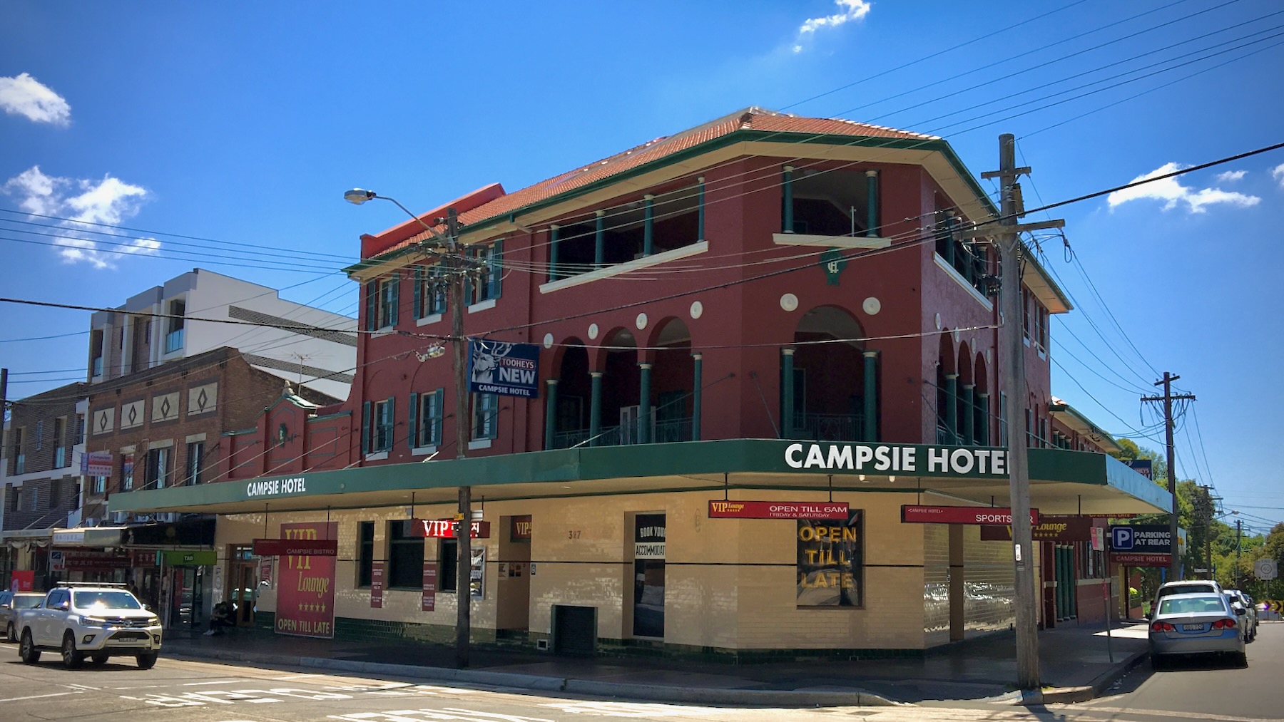 On a bright sunny day with blue sky stands a red-brick hotel, three storeys high, with the ground floor finished in cream tiles. The awning is dark green, and reads “Campsie Hotel” in white.