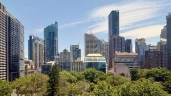 The view across Hyde Park, Sydney, from College Street. In the lower third of the image are the tops of various kinds of trees. But the main part of the image is the cityscape of the Sydney CBD, with a jumble of various skyscrapers modern and 20th century, behind the park and on the left of it. All is set against a bright blue sky and a few cirrus clouds.