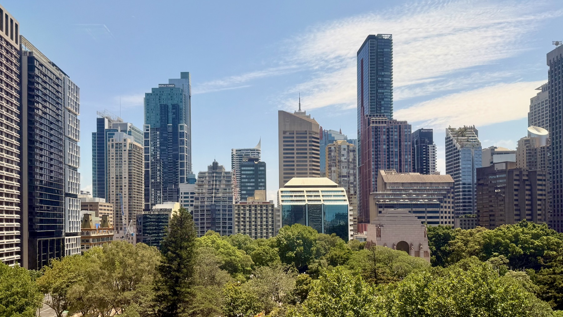 The view across Hyde Park, Sydney, from College Street. In the lower third of the image are the tops of various kinds of trees. But the main part of the image is the cityscape of the Sydney CBD, with a jumble of various skyscrapers modern and 20th century, behind the park and on the left of it. All is set against a bright blue sky and a few cirrus clouds.