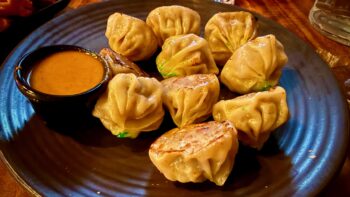 Ten little dumplings, fried, sit on a brown plate along with a small brown bowl of a brown sauce. The plate sits on a dark wooden table, and in the background is a glass of water.