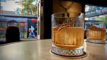 On a clean pub table sits a rocks glass, with a second one behind it. Both contain an orange-brown drink and a big fat ice cube. Behind all that is a large open window with a view of a passing tram. And on the right, through the glass door, can be seen the sign “Charlie Chan’s Bar.