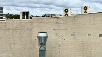 We’re looking out a window. At least two-thirds of the view is of a brick wall painted cream, heavily cracked. Rising up in the middle of the image is the shiny steel chimney of a restaurant kitchen extractor fan. Above the roofline of this wall are several air conditioning units on the roof of the next building and, in the far distance, trees and the upper parts of buildings. The sky is grey and cloudy.