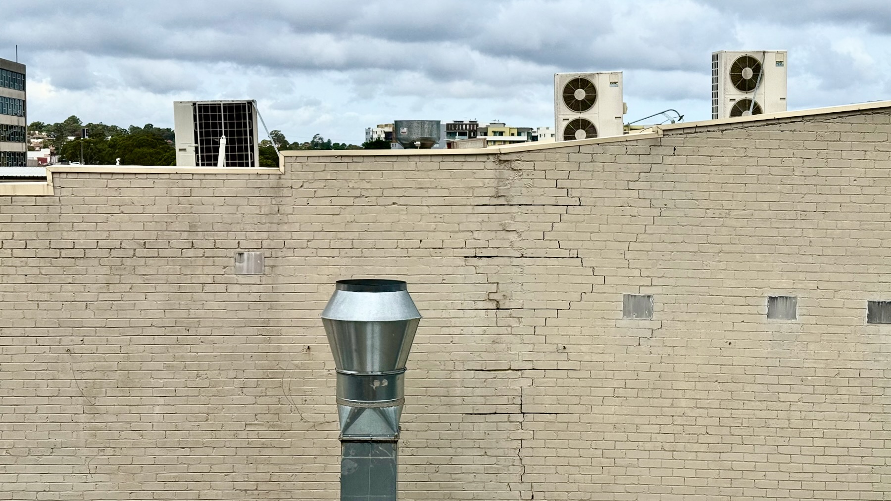 We’re looking out a window. At least two-thirds of the view is of a brick wall painted cream, heavily cracked. Rising up in the middle of the image is the shiny steel chimney of a restaurant kitchen extractor fan. Above the roofline of this wall are several air conditioning units on the roof of the next building and, in the far distance, trees and the upper parts of buildings. The sky is grey and cloudy.