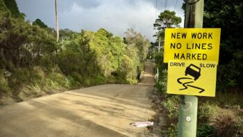A view down a rural road into a valley and then up the other side. The road has been stripped back to its foundations and surfaced with freshly-packed earth and such, ready for the next layer and, eventually, asphalt. On the right, two black on yellow signs read “NEW WORK NO LINES MARKED” and “DRIVE SLOW”. Trees and bushes line both sides of the road, with those on the left caught by the morning light.