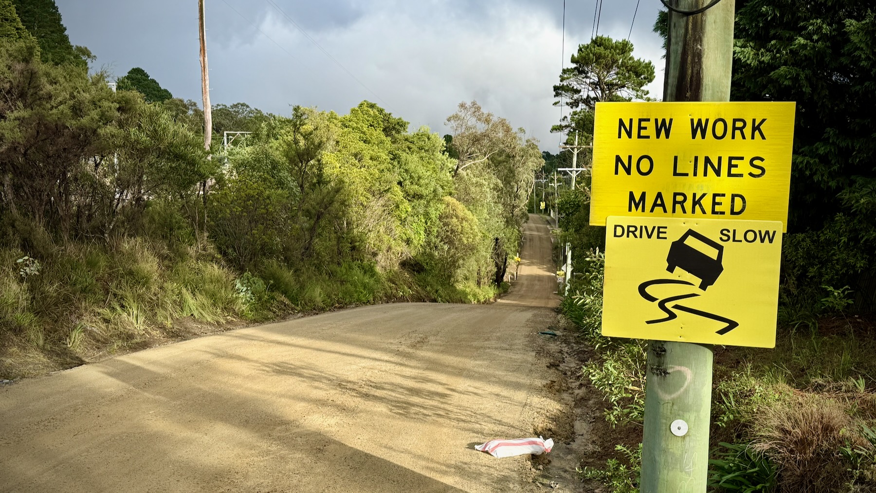 A view down a rural road into a valley and then up the other side. The road has been stripped back to its foundations and surfaced with freshly-packed earth and such, ready for the next layer and, eventually, asphalt. On the right, two black on yellow signs read “NEW WORK NO LINES MARKED” and “DRIVE SLOW”. Trees and bushes line both sides of the road, with those on the left caught by the morning light.