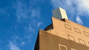 Looking up into a bright blue sky with scattered traces of cloud. A multi-storey brick apartment building looms, its side devoid of windows but decorated with square outlines of contrasting brick colours where one might expect windows to be. Tan on brown, brown on tan. At the top is a plain white box, presumably the top of the elevator shaft.