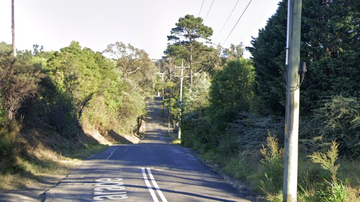 A view down a rural road into a valley and then up the other side. The road is surfaced with asphalt, but it's heavily cracked and patched, and eroding away at the sides leaving less room for traffic than there should be. Trees and bushes line both sides of the road, with those on the left caught by the morning light.