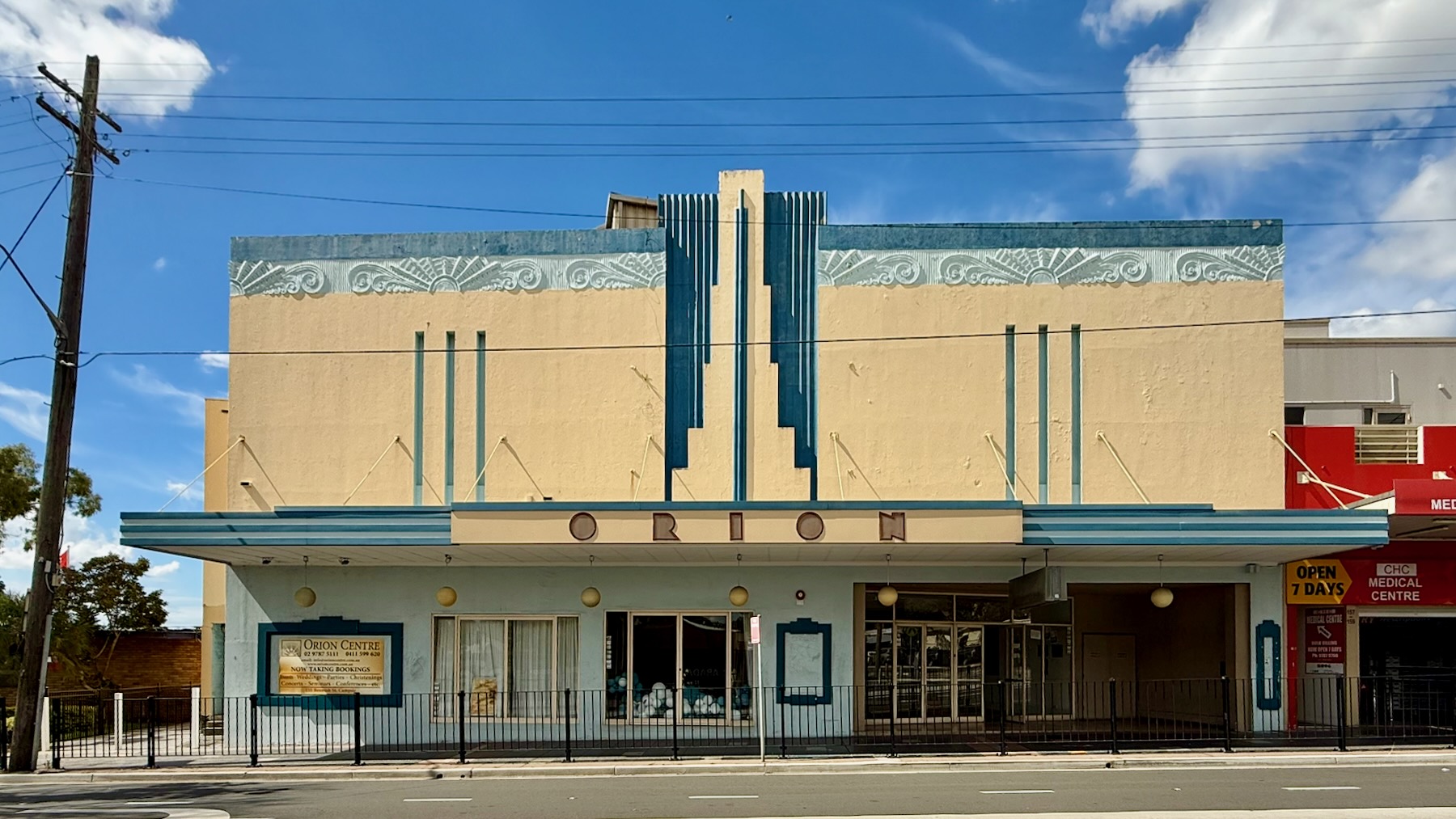 View of an art deco theatre building in cream-painted plaster and two tones of blue trim, that are the top of the building being swirly and. The marquee reads "O R I O N" in a highly stylised art deco typeface.
