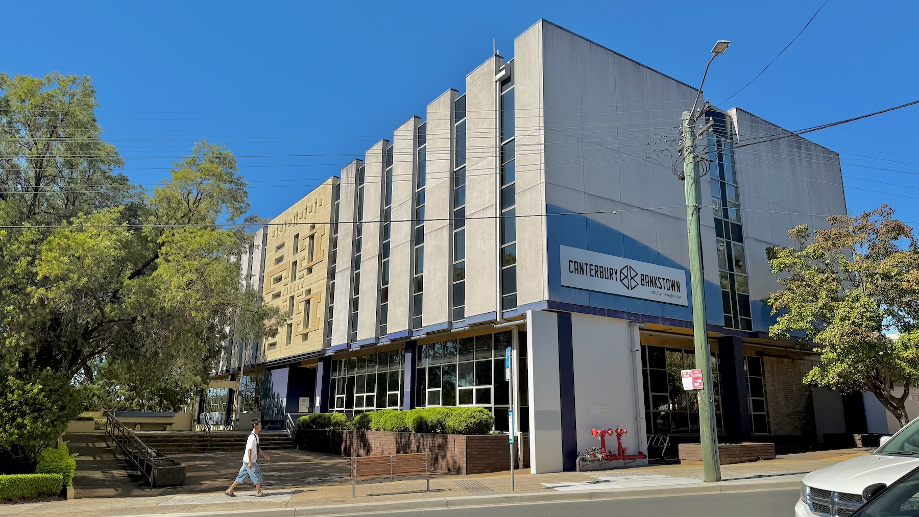 It’s a sunny day with a bright blue sky. We’re looking at a modernist office building, three storeys. It’s mostly grey concrete, with alternating vertical concrete panels and glass panels. The centre section has pale yellow panels with rectangular geometric designs. On the street side is the logo of the City of Canterbury-Bankstown. On the left are the trees of Lofts Garden, and there’s another tree on the right.