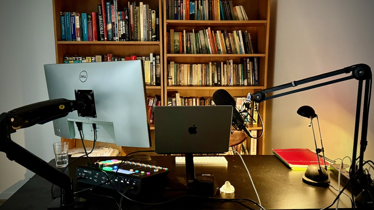 Photo of the corner of a white-walled room. In the foreground is a black desk on which sits, left to right, a RØDECaster Pro mixer, a Dell monitor, a MacBook Pro, a tiny glass Buddha, a small stack of notebooks, a desk lamp, and a mic boom holding a RØDE Procaster microphone. Behind the desk are two tall wooden bookshelves stuffed with books at random.
