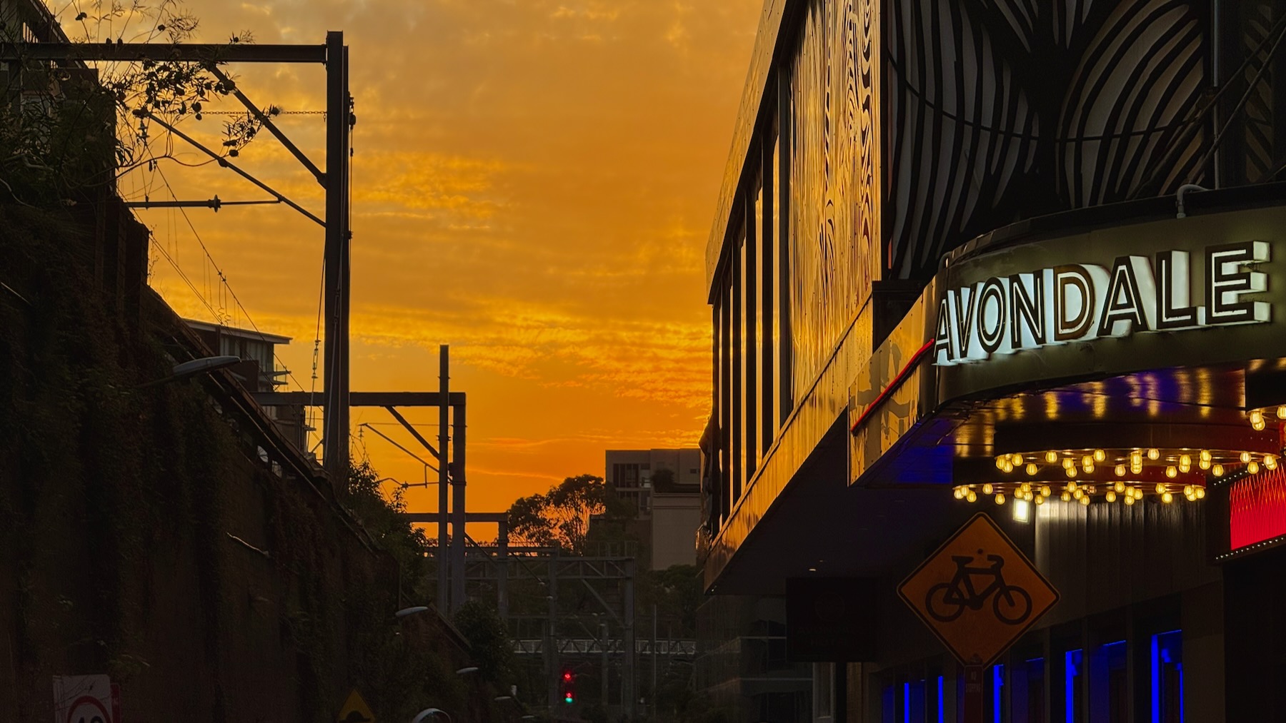 Looking down a narrow street between a towering railway embankment on the left and a pub on the right. It’s just after sunset, and the sky is a deep orange thanks to hazard reduction burns out west. The pub bears an illuminated sign, “Avondale”, and there’s naked bulbs under the small verandah.