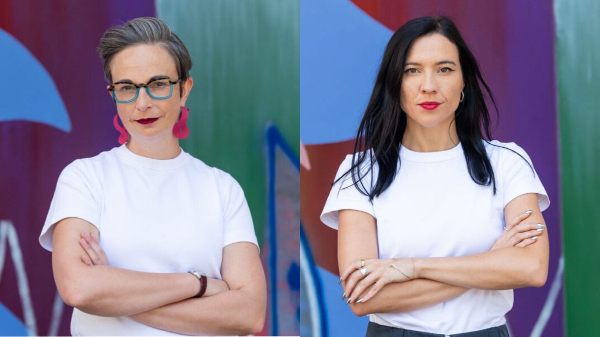 Two women stand looking into the camera. Both are wearing white t-shirts. Both have their arms folded. On the left is Johanna Weaver, with brushed-up greying hair, bright pink earrings, and glasses. On the right is Zoe Hawkins, with long straight black hair worn loose. Both are trying to look more serious than they actually are.